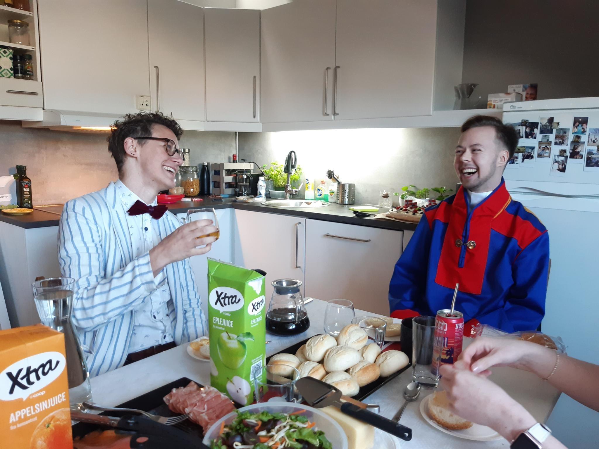 Two people laugh together over breakfast on 17 May. One wears a suit and the other a Sami gákti. Food and Norwegian flags decorate the table.