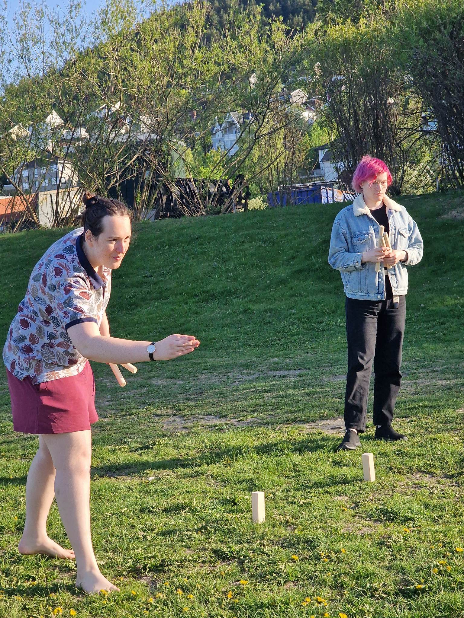 Two people play kubb on the grass. One is about to throw a wooden baton.