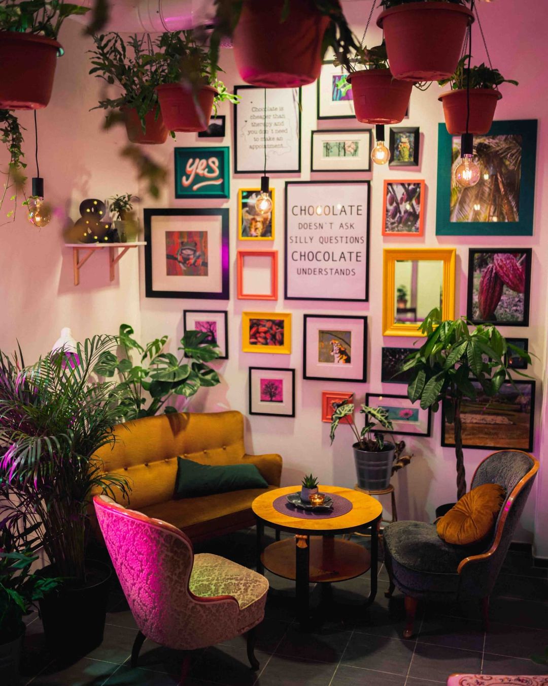 A small cafe table with three comfortable seats, hanging plants, and colourful framed pictures on the wall.