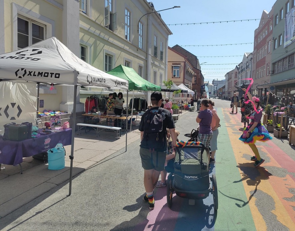 People walk between market stalls on the rainbow-painted street. A clown performs among the pride flags.