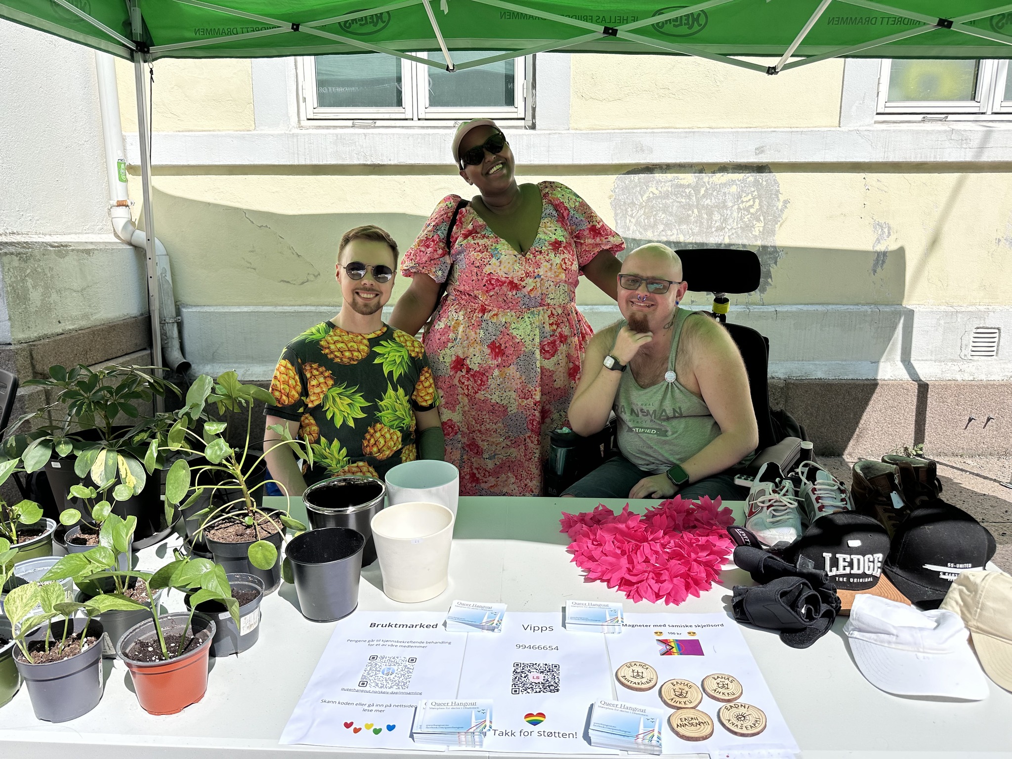 Three people smile behind a stall selling plants, handmade fridge magnets, and second-hand clothes. Pride flags and thank-you signs decorate the table.